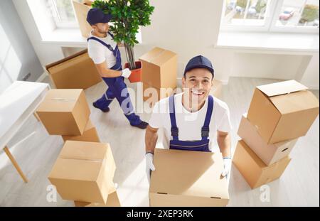 Glücklicher Angestellter, der den Service umzieht, steht in einem neuen Haus mit Pappkarton. Stockfoto