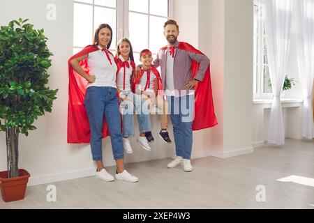 Glückliche Familie in Superheldenkostümen, die zu Hause auf der Fensterbank sitzt und in die Kamera blickt. Stockfoto