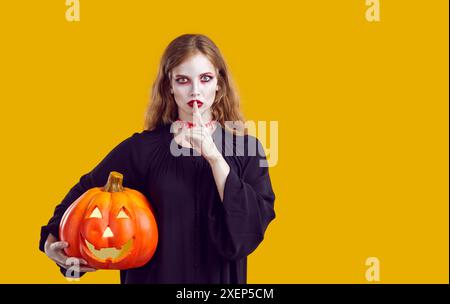 Attraktive Frau in halloween Clownschminke hält kurvenreichen Kürbis mit der „Keep quite“-Geste. Stockfoto
