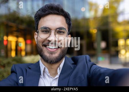 Geschäftsmann lächelt und nimmt Selfie mit Brille und Anzug im Freien mit verschwommenem Hintergrund, der modernes Gebäude und Grün zeigt Stockfoto