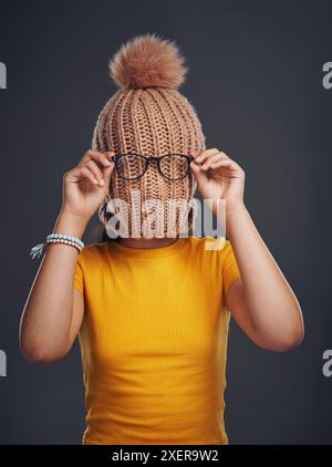 Style, Versteck und Mädchen mit Mütze im Studio mit Brille für lustige, Comic oder alberne Witze. Goofy, Mode und Frau bedecken das Gesicht mit Strickmütze und Stockfoto