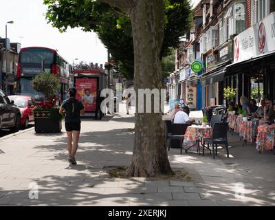 Die Leute sitzen vor Cafés und Bars in der Sommersonne in der Station Road, während Jogger vorbeifahren, North Chingford, London mit einem roten Doppeldeckerbus mit Chingford Station vor der Tür Stockfoto