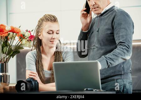 Junger Fotograf, der einem Kunden Bilder auf einem Laptop über das Telefon zeigt. Sie arbeiten im Büro des Fotografen Stockfoto