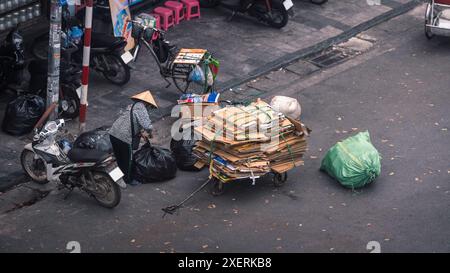 Aus der Vogelperspektive einer vietnamesischen Frau, die einen traditionellen asiatischen konischen Strohhut trägt, sammelt Pappe und Müll für das Recycling entlang einer Straße in Hanoi Old Stockfoto