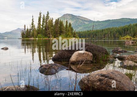 Ein horizontales Foto von kontrastfarbenen Felsen in Lake Wenatchee, Washington, mit einer kleinen Insel aus Bäumen und Cascade Mountains im Hintergrund. Stockfoto