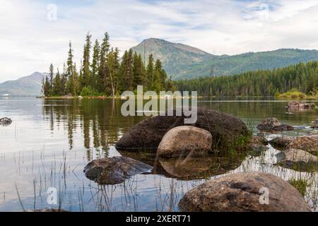 Ein horizontales Foto von kontrastfarbenen Felsen in Lake Wenatchee, Washington, mit einer kleinen Insel aus Bäumen und Cascade Mountains im Hintergrund. Stockfoto