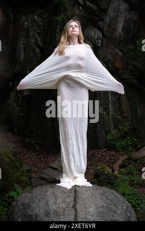 Die Frau steht auf einem Felsen, bedeckt in einem fließenden weißen Stoff, ihre Arme gestreckt. Sie blickt nach oben und schafft eine ätherische und stärkende Atmosphäre für Agi Stockfoto