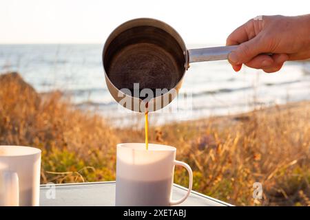 Ein Reisender gießt vor dem Hintergrund des Meeres gebrühten Kaffee in Tassen. Freizeitangebote im Freien. Reisen und Tourismus. Stockfoto