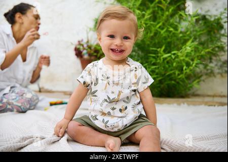 Authentisches Porträt eines niedlichen Jungen, der in die Kamera blickt und draußen im Garten auf einer Decke sitzt. Fröhliche Frau in weißem Hemd liebevoll Stockfoto