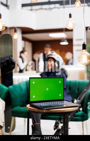 Detaillierte Aufnahme eines digitalen Geräts mit isolierter Copyspace-Vorlage auf der Bank im Loungebereich des luxuriösen Skiresorts. Frau in Wintersportausrüstung vor dem Hintergrund eines Laptops mit grünem Bildschirm. Stockfoto