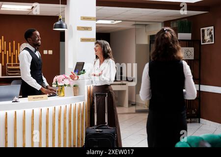 Der freundliche afroamerikanische Concierge begrüßt ältere Frauen im Ruhestand und hilft beim Check-in. Im Luxusresort spricht eine Seniorin mit männlichen Rezeptionisten über die besten Urlaubsaktivitäten. Stockfoto