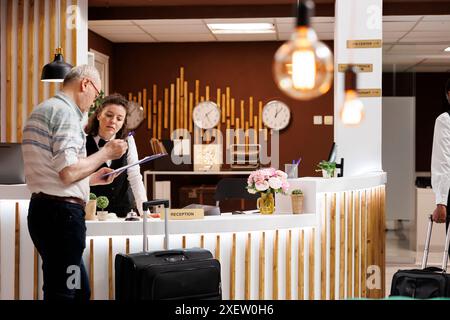 Senior männliche Touristen checken an der Hotelrezeption ein, bereit für einen lustigen Urlaub. Der freundliche weibliche Concierge hilft älteren Reisenden, während er Reservierungsformulare ausfüllt. Stockfoto