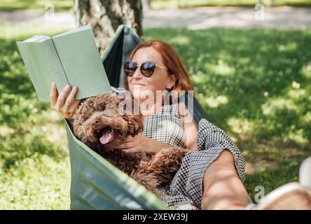 Frau, die Buch liest, entspannt sich in der Hängematte mit ihrem flauschigen braunen Maltipoo-Hund an sonnigem Tag. Sowohl zufrieden als auch glücklich. Diese Outdoor-Szene fängt Jo ein Stockfoto