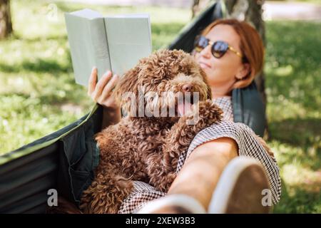 Frau, die Buch liest, entspannt sich in der Hängematte mit ihrem flauschigen braunen Maltipoo-Hund an sonnigem Tag. Sowohl zufrieden als auch glücklich. Diese Outdoor-Szene fängt Jo ein Stockfoto