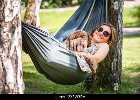 Frau entspannt sich in der Hängematte mit ihrem flauschigen braunen Maltipoo Hund an sonnigen Tagen. Sowohl zufrieden als auch glücklich. Diese ruhige Outdoor-Szene fängt die Freude von bo ein Stockfoto