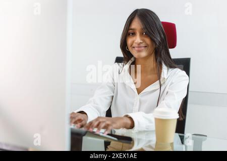 Eine junge Geschäftsfrau, die an ihrem Schreibtisch in einem modernen Büro arbeitet. Professionelle Frau lächelt, während sie einen Computer benutzt. Positives und produktives Arbeitsumfeld Stockfoto