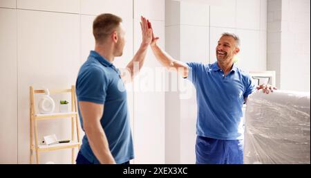 Professionelle Mover in Uniform und Team, die eine hohe Geste von fünf geben, nachdem sie eine weiße Matratze an einen zufriedenen Kunden geliefert haben. Stockfoto