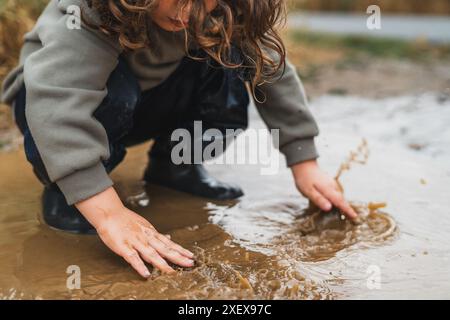 Ein Kind spielt im Regen und spritzt Wasser mit den Händen. Die Szene ist verspielt und fröhlich, während das Kind die nassen und schlammigen Bedingungen genießt Stockfoto