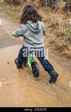 Ein kleiner Junge fährt mit einem grünen Fahrrad durch einen schlammigen Pfad. Der Junge trägt einen grauen Hoodie und eine blaue Hose. Die Szene ist nass und schlammig, und der Junge ich Stockfoto