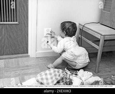 Gefährlich in den 1960ern Ein Kind sitzt auf dem Boden und ist neugierig auf die Steckdose in der Wand. Sie hält einen Stecker in der einen Hand und fesselt die Steckdose mit der anderen. Damals hatten elektrische Stecker nicht den Schutz, den moderne Stecker haben. Unfälle ereigneten sich, als Kinder einen Stromschlag erlitten hatten. Schweden 1960er Jahre Stockfoto