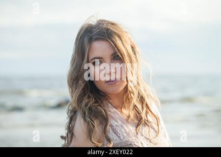 Wunderschöne junge Erwachsene Frau mit natürlichem Make-up und langen welligen braunen Haaren auf blauem Meer Strand Outdoor Porträt Stockfoto