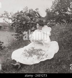 Vintage-Foto der Liebe. Er liebt mich, er liebt mich nicht. 1906 Frau saß auf dem Boden und pflückte Blütenblätter aus Blumen, während der Mann hinter dem Busch zuschaut. Stockfoto