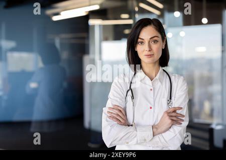 Selbstbewusste Ärztin mit Stethoskop im modernen Krankenhausraum. Junge professionelle Pose mit überkreuzten Armen, zeigt Autorität und Vertrauenswürdigkeit in der Medizin, im Gesundheitswesen Setting Stockfoto