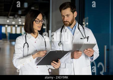 Ärzte stehen im Krankenhauskorridor und diskutieren die Krankenakte des Patienten. Männlicher Arzt mit Tablette, weiblicher Arzt mit Klemmbrett. Medizinische Fachkräfte engagierten sich für die kollaborative Gesundheitsbeurteilung. Stockfoto