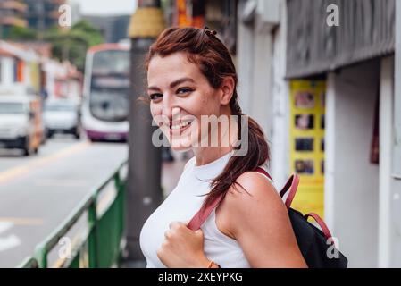 Eine fröhliche junge Frau mit Rucksack lächelt, während sie auf einer belebten Straße steht und eine lebhafte urbane Umgebung präsentiert. Stockfoto
