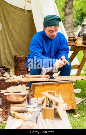 Mittelalterliches Lager, ein Reenactor in historischen Kostümen, der Holz formt, mit einem kleinen Handmesser und einem kleinen Tisch mit Werkzeugen vor ihm. Stockfoto