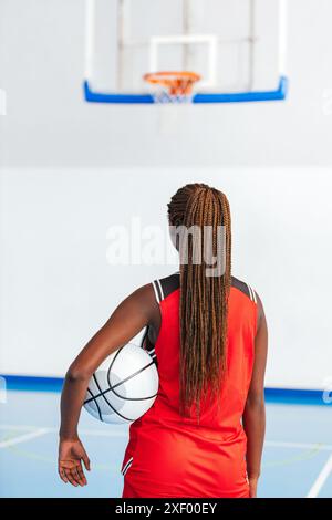 Foto: Eine Basketballspielerin in roter Uniform steht auf einem Hallenplatz, hält einen Basketball und fokussiert auf den Korb, bereit für einen Schuss. Sie Stockfoto