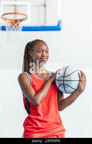 Vertikale Foto junge Basketballspielerin in roter Uniform hält einen Basketball nahe an ihre Brust, während sie auf einem Hallenplatz steht und foc zeigt Stockfoto