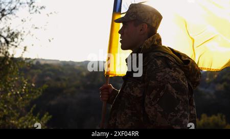 Soldat der ukrainischen Armee steht mit einem blau-gelben Banner auf dem Feld. Männliches Militär in Tarnuniform hält eine winkende Flagge der Ukraine. Sieg Stockfoto