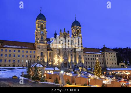 Einsiedeln, Schweiz - 10. Dezember 2023: Das Benediktinerabtei Einsiedeln mit seiner mächtigen Basilika in blauer Stunde und Weihnachtsmarkt im Fro Stockfoto