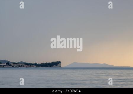 Ruhiges Meer bei Sonnenuntergang im späten Frühling. Resort in Roda und Klippen daneben. Bewölkter Himmel. Insel Othonoi am Horizont. Acharavi, Korfu, Griechenland. Stockfoto