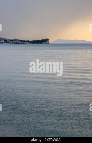 Ruhiges Meer bei Sonnenuntergang im späten Frühling. Resort in Roda und Klippen daneben. Bewölkter Himmel. Insel Othonoi am Horizont. Acharavi, Korfu, Griechenland. Stockfoto