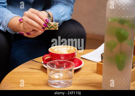 Frau, die Zucker aus einer kleinen Packung in Espresso auf einem hölzernen Kaffeetisch gießt. Nahaufnahme der Kaffeezubereitung. Stockfoto