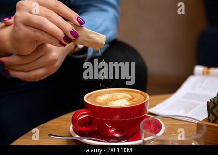 Frau, die Zucker aus einer kleinen Packung in ihren Kaffee auf einem Kaffeetisch gießt. Nahaufnahme. Stockfoto