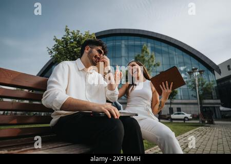 Junge Berufstätige, die auf einer Bank vor dem modernen Bürogebäude lachen und arbeiten Stockfoto