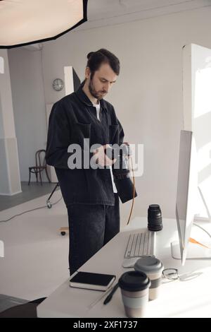 Vertikales Porträt eines bärtigen, männlichen Fotografen, der die Kamera hält und schwarz im kontrastweißen Fotostudio trägt Stockfoto