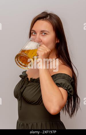 Eine Frau mit langen braunen Haaren in einem schulterfreien grünen Kleid, lächelt und trinkt Bier aus einem großen Glas. Stockfoto