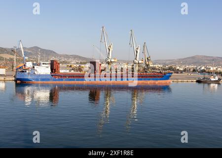 Ein großes Frachtschiff legte an einem geschäftigen Industriehafen an, mit Kränen für den Frachtumschlag und einer Stadtlandschaft im Hintergrund. Stockfoto