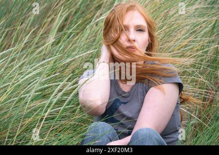 Rothaarige Frau sitzt in hohem, windgepeitschten Gras, ihr Haar bläst sanft über ihr Gesicht. Blickt nachdenklich in die Ferne und fängt ein ruhiges Kontem ein Stockfoto