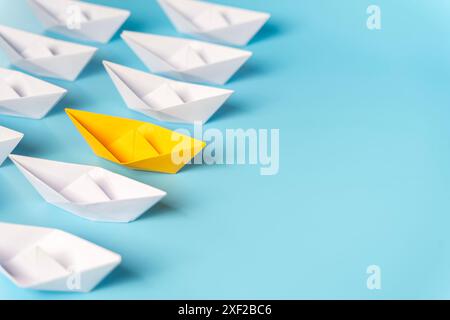 Gelbes Papierschiff, das vor anderen Weißpapierschiffen auf blauem Wasser segelt. Gelbes Papierboot, das auf hellem Hintergrund von anderen wegschwebt, flache Lay wi Stockfoto