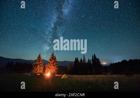 Nachtcampen in den Bergen unter Sternenhimmel. Touristenzelte auf dem Campingplatz unter dem Himmel voller Sterne mit Milchstraße. Silhouette von Wanderer, die sich am brennenden Lagerfeuer ausruhen. Tourismuskonzept, Abenteuer. Stockfoto