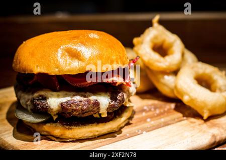 Hamburger mit Fleisch, Käse und Speck auf einem Brötchen mit Zwiebelringen auf einem Holzteller im Pub Stockfoto