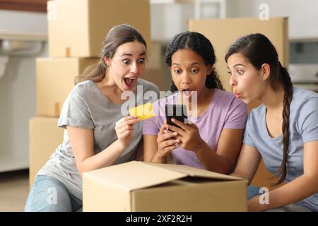 Drei verblüffte Mitbewohner, die online einkaufen, mit Kreditkarte, die das Handy überprüft, ziehen aus Stockfoto