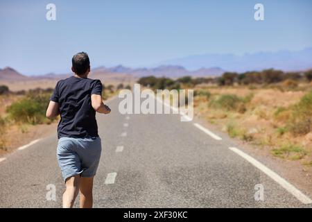 Ein Reisender hebt seine Arme zum Sieg, während er auf einer endlosen Straße durch die marokkanische Wüste geht, die Freiheit und Abenteuer symbolisiert. Stockfoto