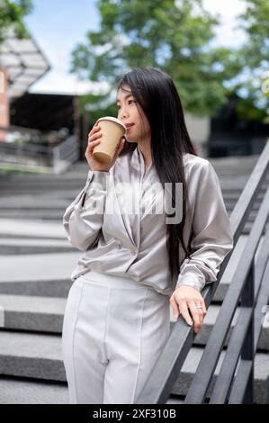 Eine attraktive asiatische Geschäftsfrau in stilvoller Geschäftskleidung trinkt ihren Kaffee, während sie auf der Treppe im Freien in der Stadt steht. Modernes Stadtleben Stockfoto