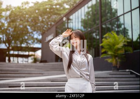 Eine attraktive asiatische Geschäftsfrau in stilvoller Bürokleidung schirmt ihr Gesicht vor der Sonne ab, während sie in der Stadt auf einem die Treppe hinunter geht Stockfoto